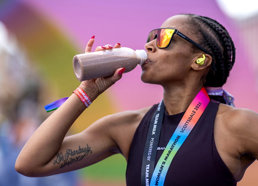 Athlete wearing sunglasses drinks a chocolate protein shake after a race, with a marathon medal around her neck.